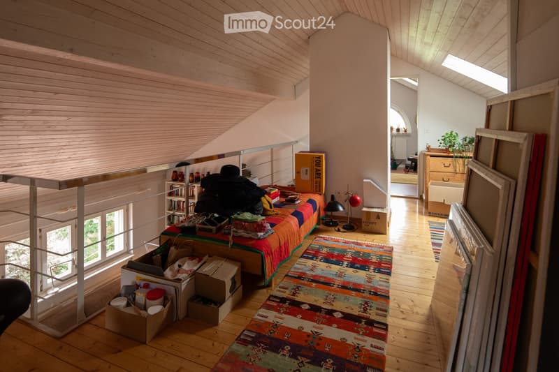 A cozy, loft-style bedroom with wooden floors and slanted ceilings. Features a bed with a colorful quilt, a small table with a lamp, a bookshelf, and a potted plant. The room is partially unpacked with boxes and items scattered around.