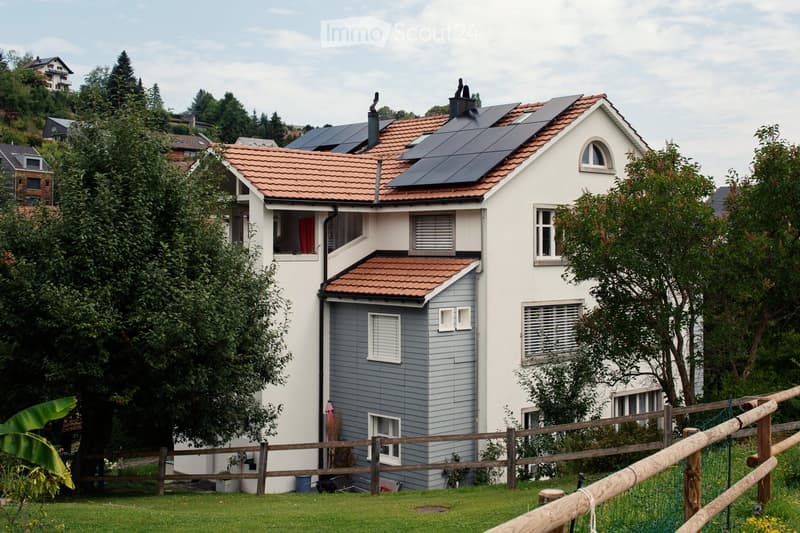 Two-story house with white and grey exterior, red-tiled roof, solar panels, multiple windows, and a wooden fence surrounding the property.