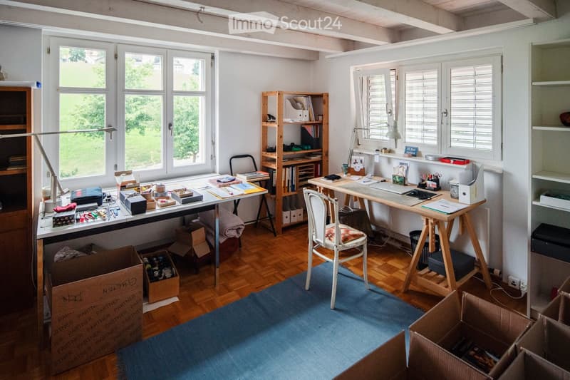 Bright, airy room with wooden flooring and large windows. Contains two desks with chairs, a wooden shelf, a lamp, and various office supplies. Several cardboard boxes are scattered around, indicating a move or organization.