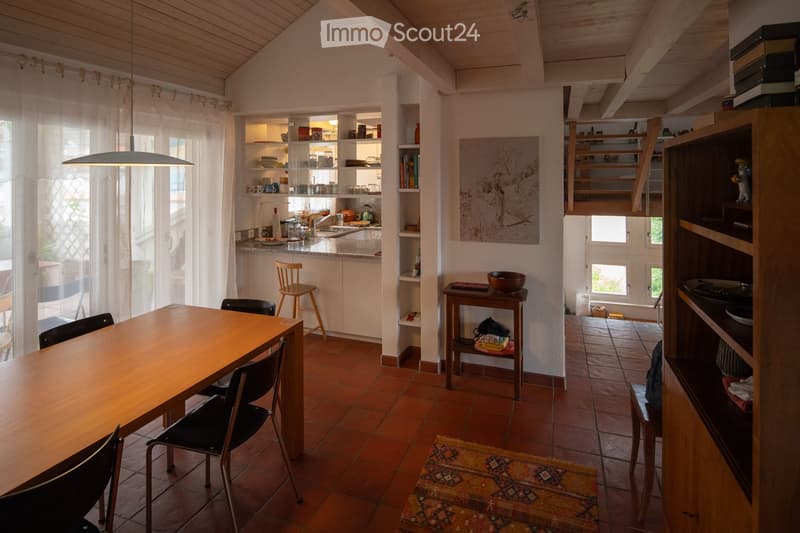 A cozy, open-plan living and dining area with wooden furniture, a kitchen counter, shelves, and a painting on the wall.