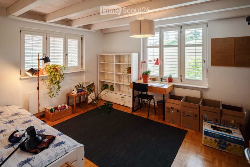 Bright bedroom with wooden flooring, large windows, a bed with a patterned duvet, a desk with a chair, a bookshelf, and several potted plants. The room also features a floor lamp, a small table with books, and several cardboard boxes.