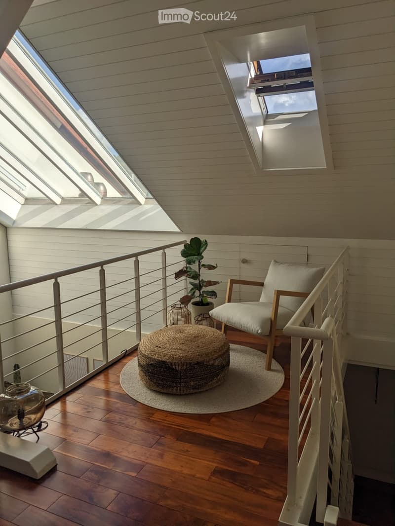 A cozy, well-lit room with wooden flooring, a modern chair, a round woven stool, a potted plant, and a decorative table lamp near the railing.