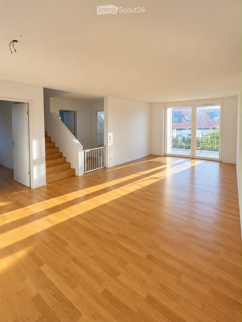 Empty room with wooden flooring, white walls, and large windows letting in natural light. A staircase leads to another level, and a baby gate is visible near the stairs.