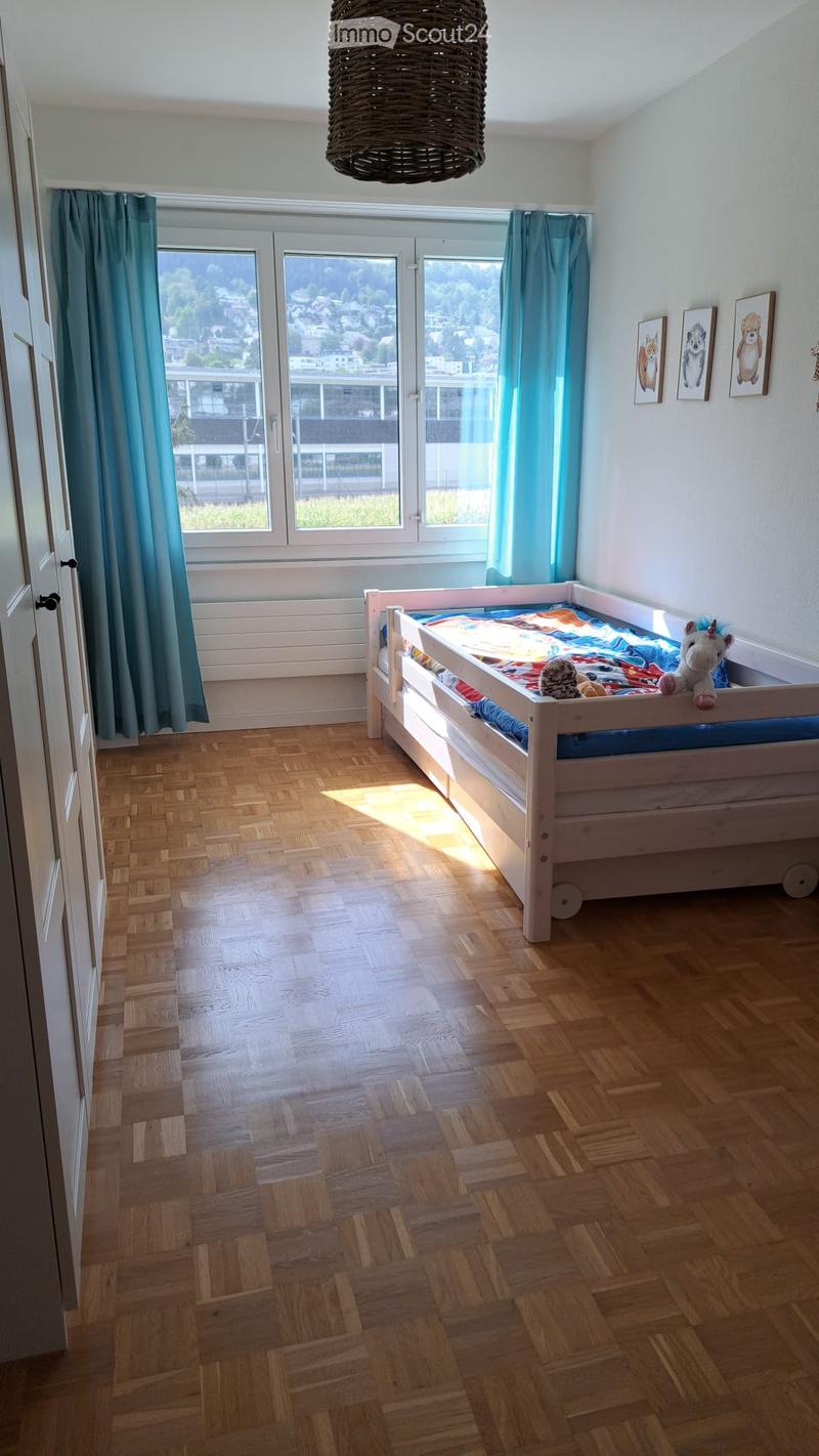 Bright bedroom with wooden flooring, a single bed with colorful bedding, a stuffed unicorn toy, and framed pictures on the wall. Large windows offer a scenic view of the surroundings.