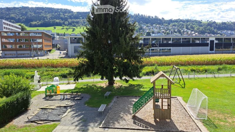 Playground area with swings, slide, and benches, surrounded by grassy fields and modern buildings in the background.