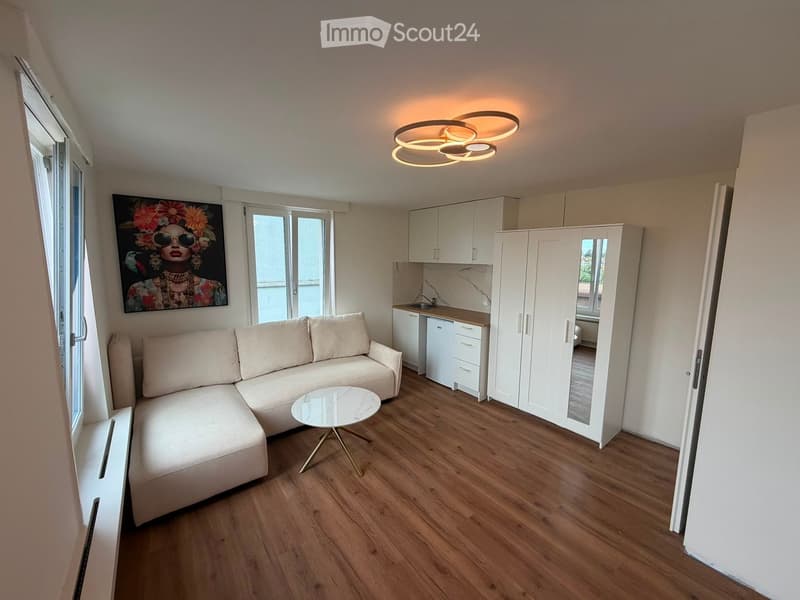 Modern, compact living space with a light-colored sectional sofa, a small round coffee table, a fitted kitchen area with cabinets and a fridge, and a large mirror on the wall. The room features wooden flooring and large windows allowing natural light.