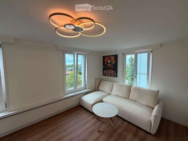 Modern living room with light-colored sofa, round marble coffee table, large windows, and a contemporary ceiling light fixture.