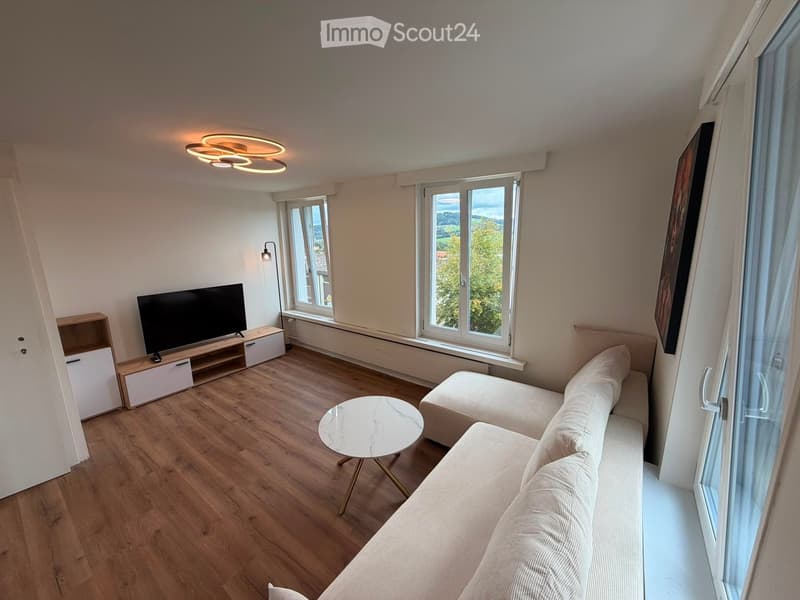 Modern living room with wooden flooring, large windows, a beige sofa, a marble coffee table, a flat-screen TV on a wooden stand, and a wall-mounted cabinet.