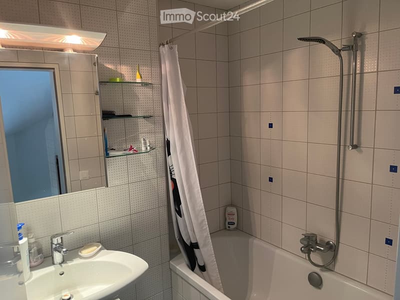 Modern bathroom with a white tiled wall, a sink with a mirror cabinet, a bathtub with a showerhead, and a shower curtain. The mirror cabinet has shelves with various toiletries.