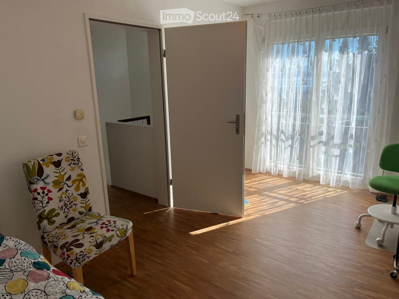A small, well-lit room with wooden flooring, a floral-patterned chair, a green office chair, and a small white stool. The room has a door leading to another space and sheer white curtains on the windows.
