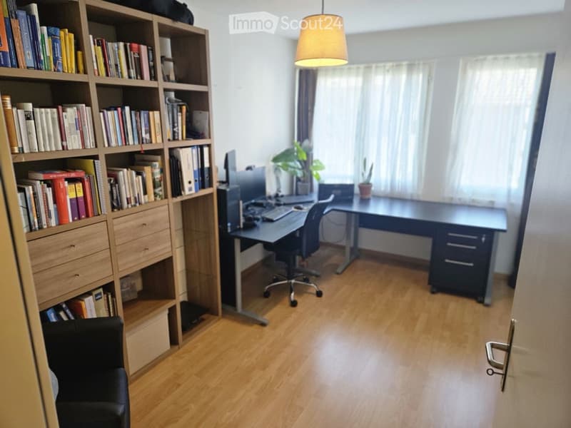 A well-lit study or home office with wooden flooring, a large bookshelf filled with books, a desk with a computer setup, and a comfortable chair.