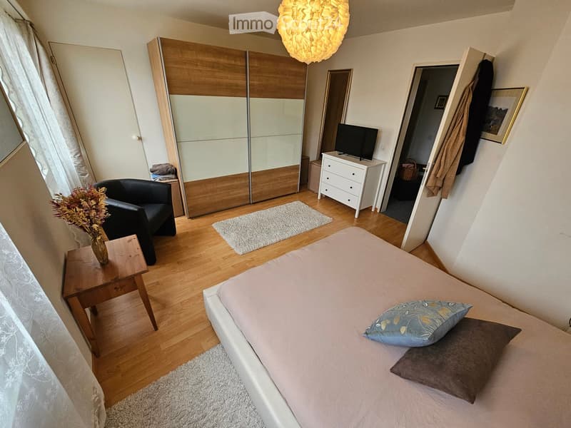 Cozy bedroom with a white bed, nightstand, wooden wardrobe, black armchair, and a small table with a vase of flowers. The room features wooden flooring and large windows with sheer curtains.