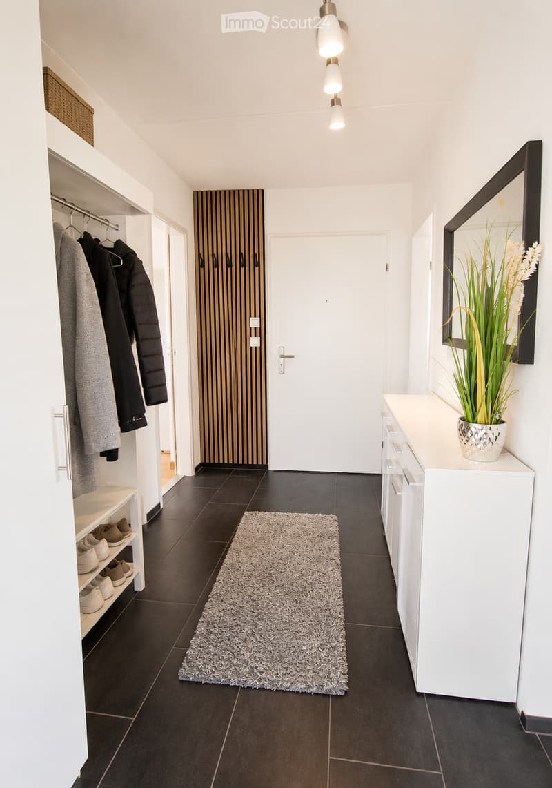 Modern hallway with dark tiled floor, white walls, built-in shoe rack, coat rack, small table with plant, and a large mirror on the wall.