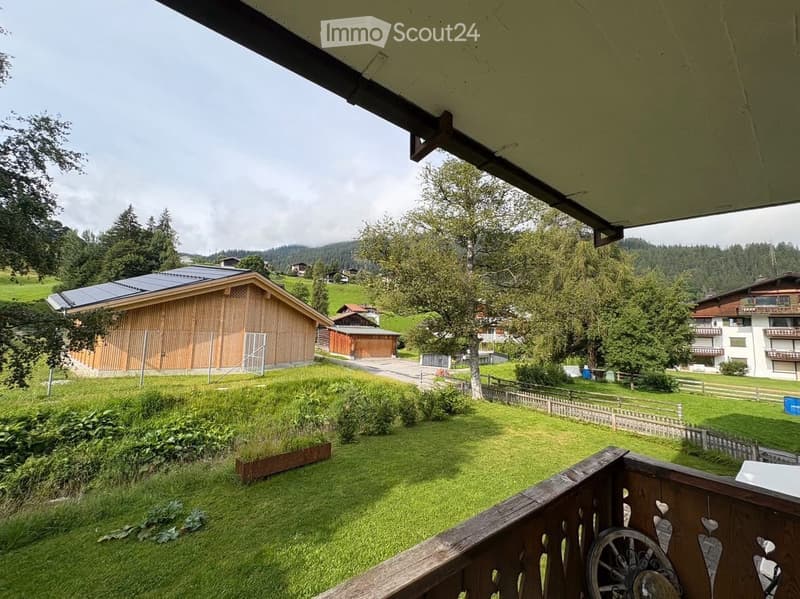 Balcony overlooking a green lawn, wooden fence, and distant houses with trees and mountains in the background.