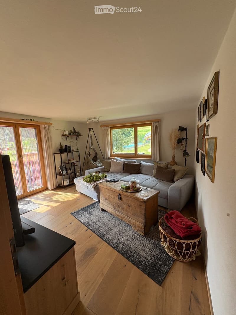 Bright living room with wooden flooring, large windows, grey sectional sofa, coffee table, wicker basket, and wall-mounted shelves.