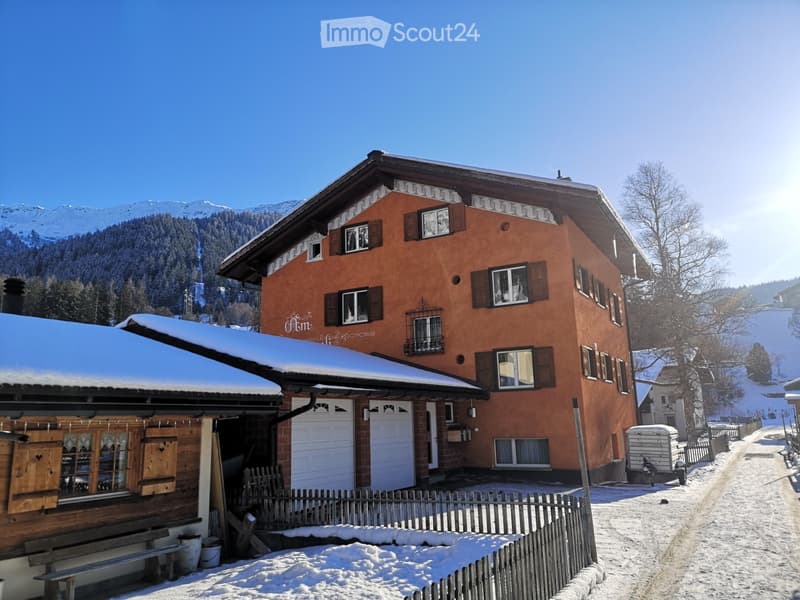 A two-story house with a red-orange facade, multiple windows, and a snow-covered roof, situated in a snowy mountain landscape.