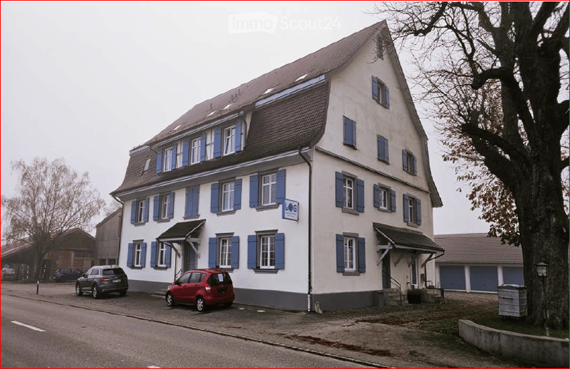 Two-story building with white walls, blue shutters, and a brown roof. Two cars are parked in front of the building.