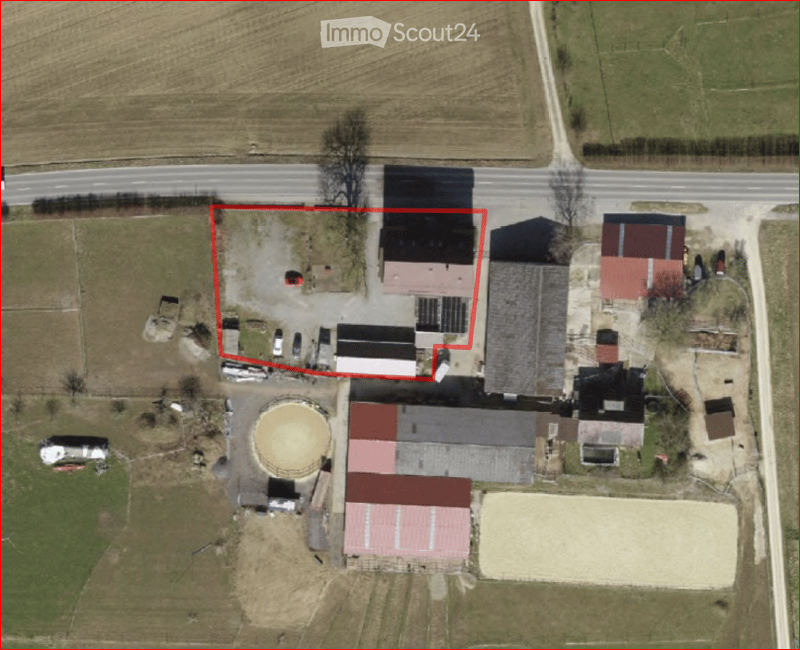 Aerial view of a rural farm complex with multiple buildings, including barns, storage sheds, and a circular silo. The main house is highlighted in red, surrounded by open fields and a road.