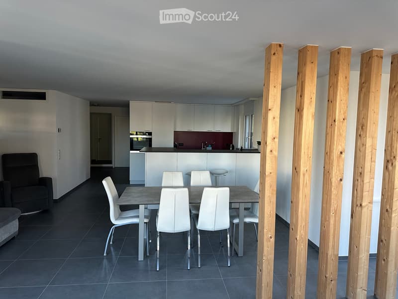 Open-plan living and dining area with a grey dining table, white chairs, a black sofa, and a modern kitchen with white cabinets and a dark countertop.