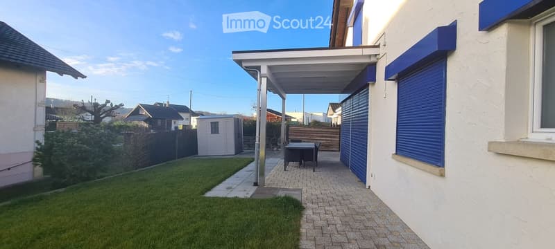 Outdoor patio area with a covered walkway, small table and chairs, lawn, and surrounding buildings.