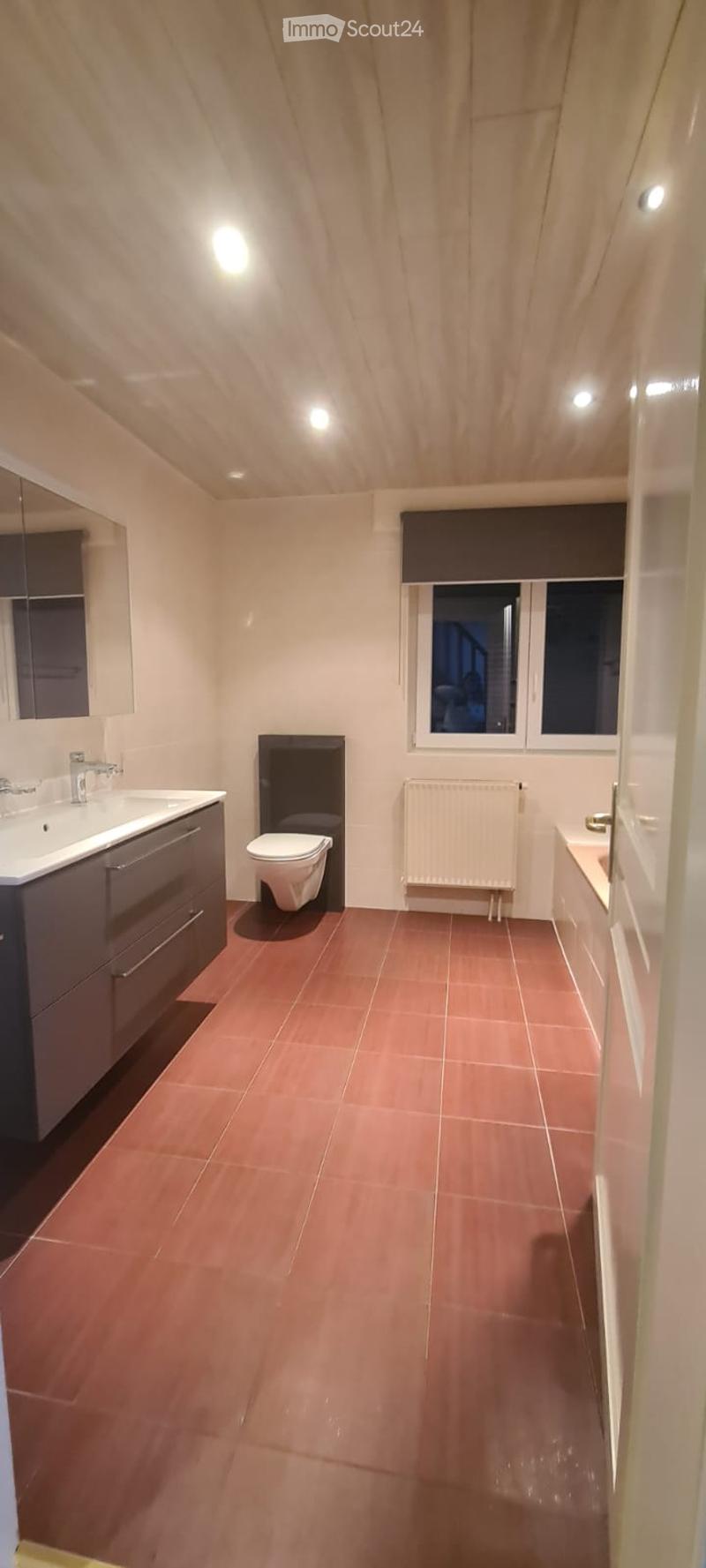 Modern bathroom with red tiled floor, white sink with faucet, toilet, radiator, and a bathtub. The room features a wooden slatted ceiling and a window with dark blinds.