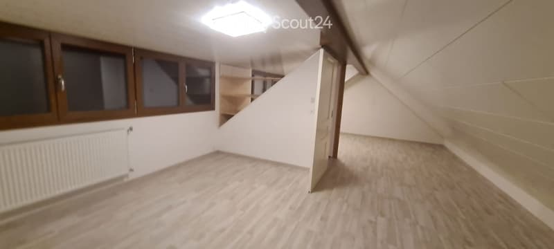Empty attic room with wooden flooring, white walls, and built-in shelving. Features a window with wooden frame and a radiator below it.