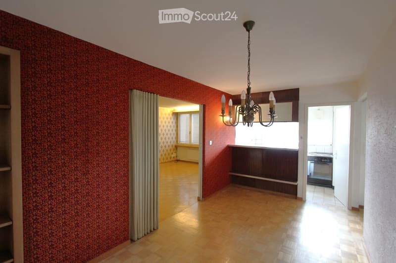 Empty room with wooden flooring, red patterned wallpaper, and a chandelier hanging from the ceiling. The room has an open doorway leading to another room with a window and a radiator.