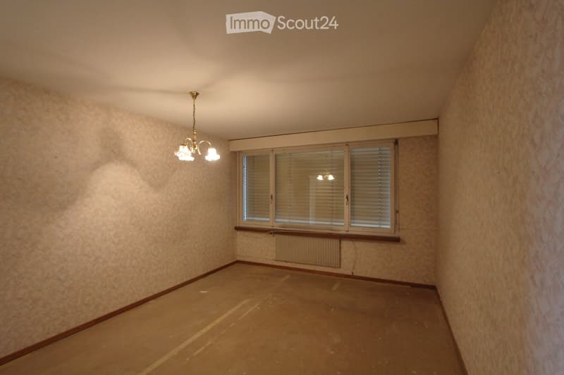 Empty room with beige walls, wooden floor, large window with blinds, radiator, and a chandelier light fixture.
