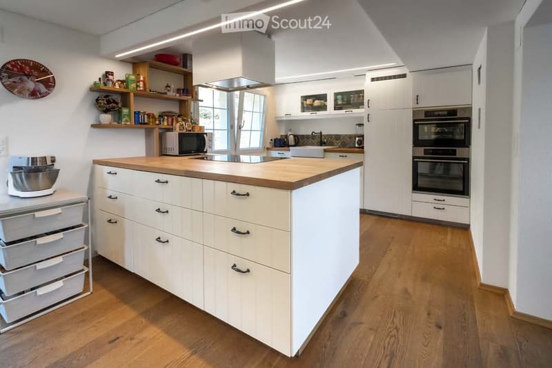 Modern kitchen with white cabinets, wooden island, microwave, oven, fridge, blender, and shelves with various items.