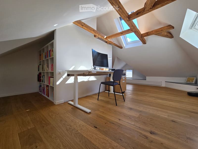 Bright attic room with wooden floors, exposed wooden beams, skylights, a white adjustable desk, a black chair, a bookshelf filled with books, and a computer screen.