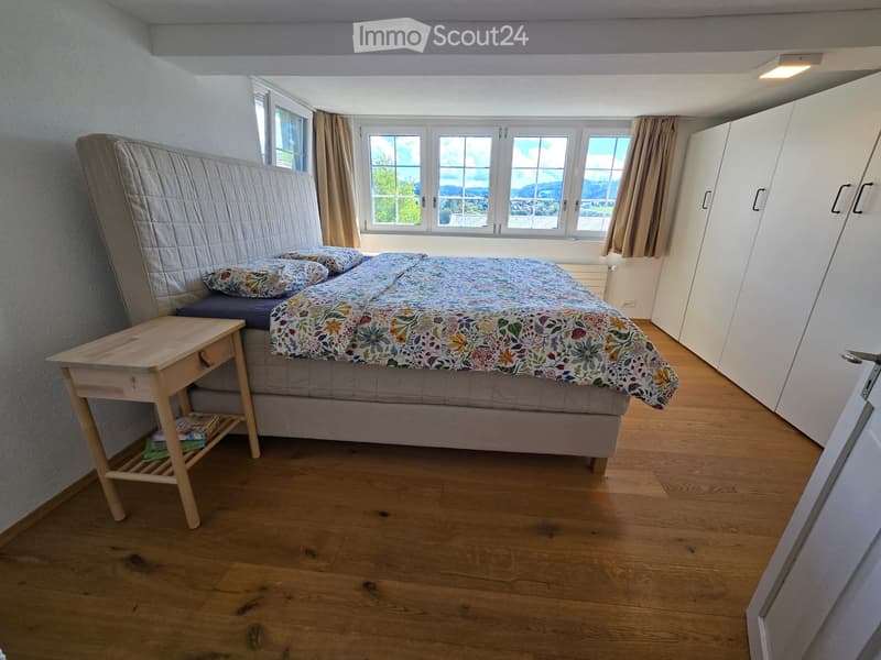 Bright bedroom with wooden flooring, large windows, a neatly made bed with floral bedding, a nightstand with books, and a built-in wardrobe.