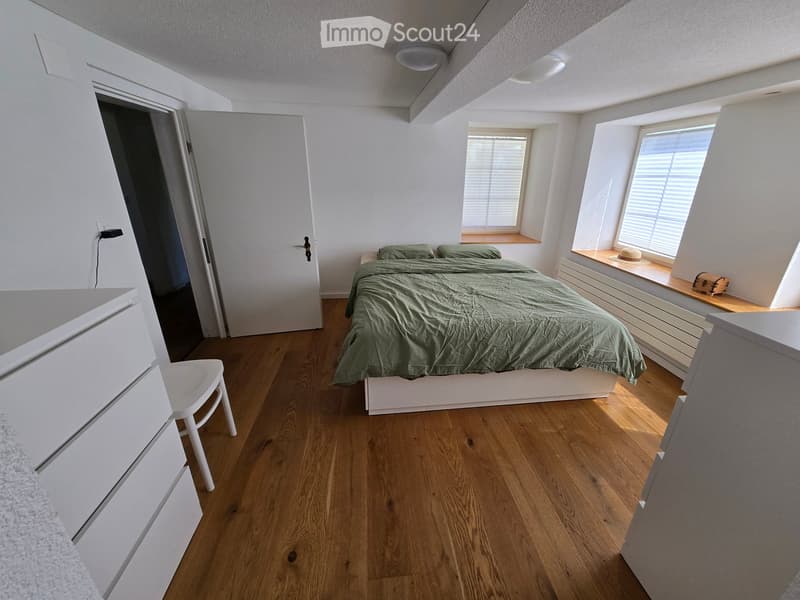 Cozy bedroom with wooden flooring, a bed with green bedding, a white chair, and a small window seat with a hat and suitcase.