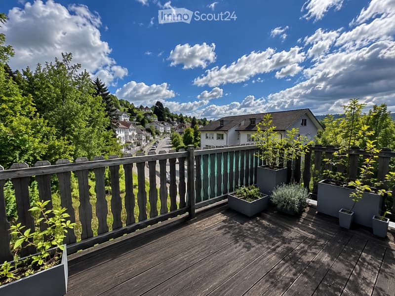 Wooden balcony with railing, potted plants, overlooking a scenic town with houses and greenery under a partly cloudy sky.