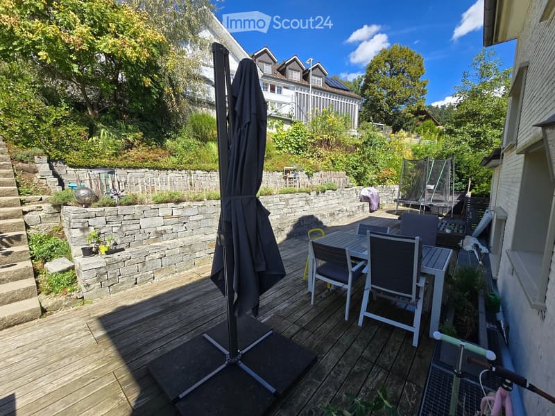Wooden deck with a table, chairs, and an umbrella, surrounded by a stone wall, greenery, and steps leading up to the house.
