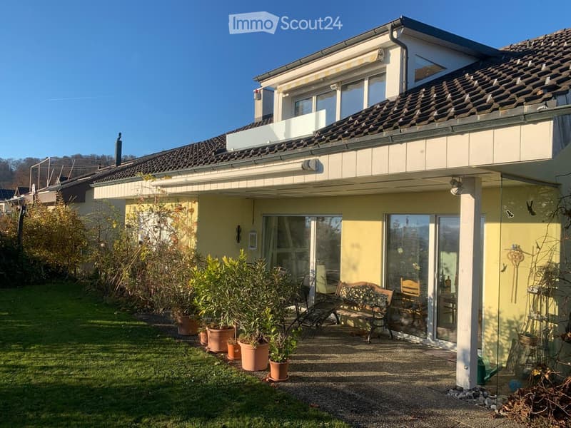 Two-story house with a tiled roof, large windows, and a balcony on the upper level. The front yard features a well-maintained lawn, several potted plants, and outdoor seating including a bench and chairs.