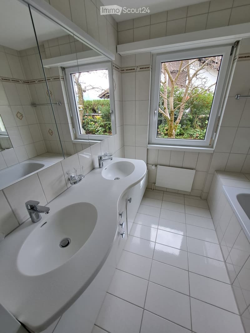 Modern bathroom with white tiles, a large sink with two faucets, a bathtub, and a large mirror. Natural light streams in through two windows showing a garden view.