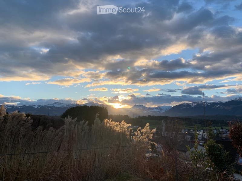 A scenic view of a town with mountains in the background during sunset, featuring tall grasses in the foreground.