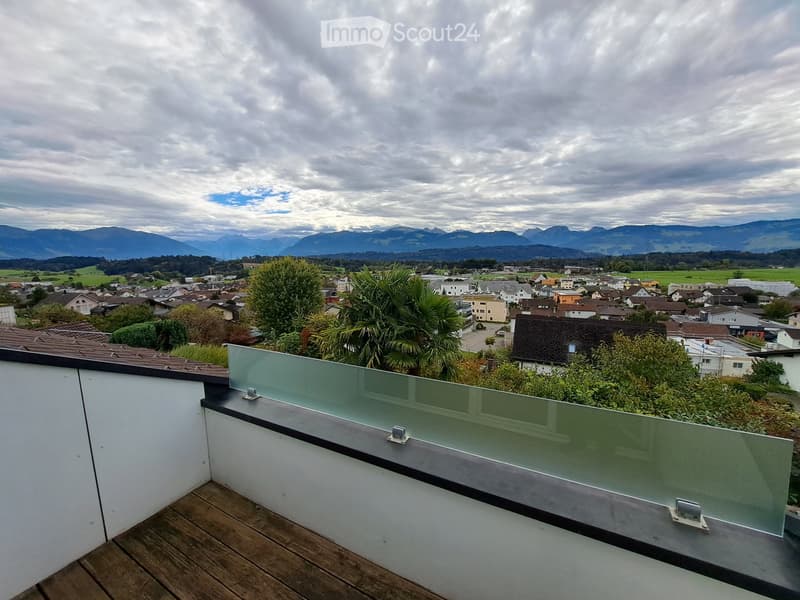 A modern rooftop terrace with wooden decking, glass railing, and scenic views of a town and distant mountains.