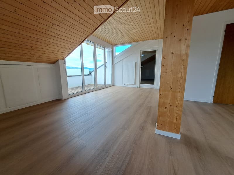 Empty room with wooden flooring, white walls, and large windows offering a view. The ceiling is slanted with wooden beams, and there is a wooden pillar in the center.