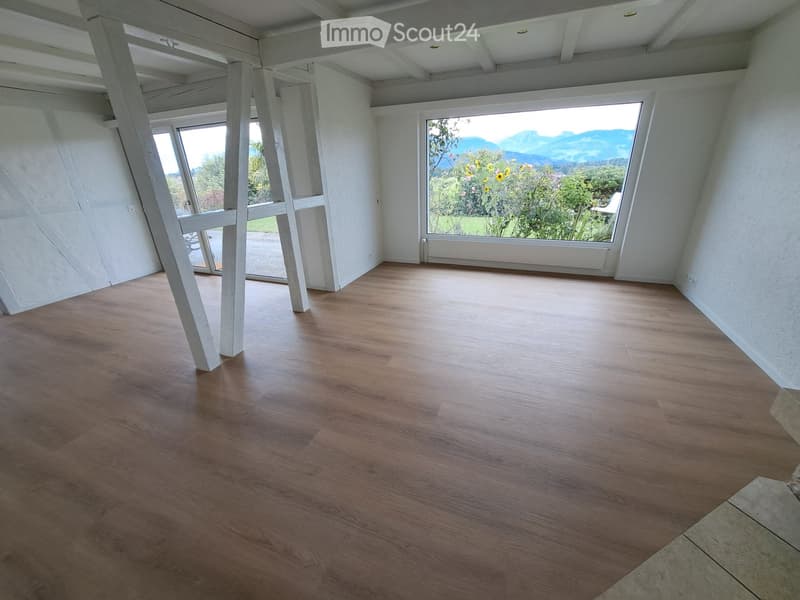 Empty room with wooden flooring, white walls, large windows, and visible beams in the ceiling.