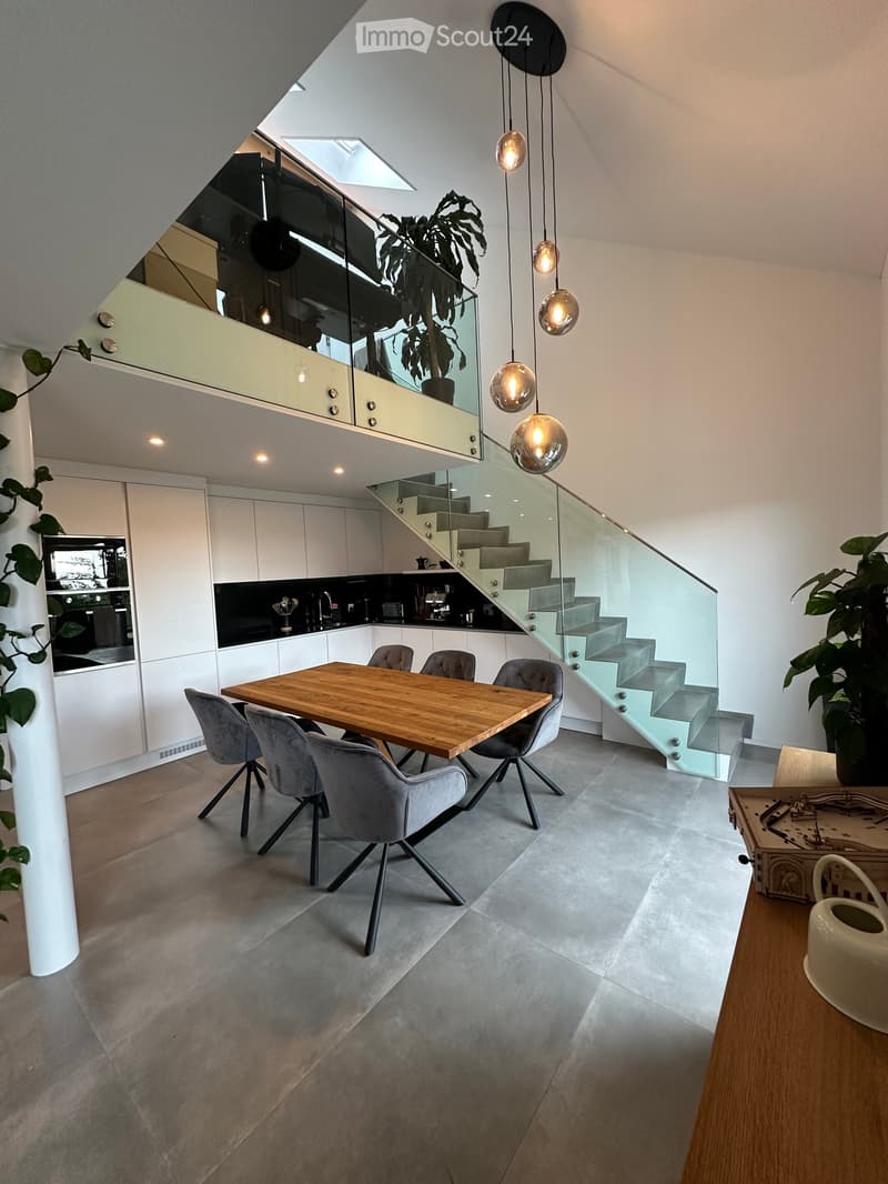 Modern open-plan dining and kitchen area with a wooden dining table, grey chairs, white cabinets, and a staircase leading to an upper level.