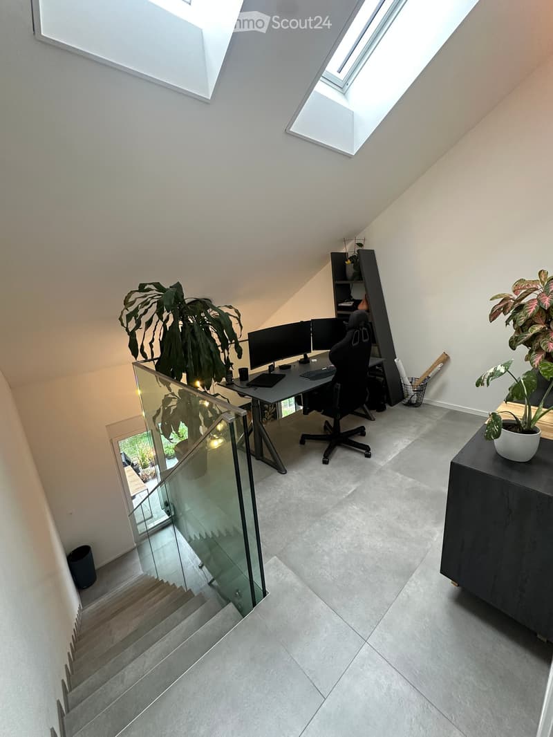 Modern, well-lit room with a sleek desk setup, two computer monitors, ergonomic chair, and large plants. Features a glass staircase leading downwards.