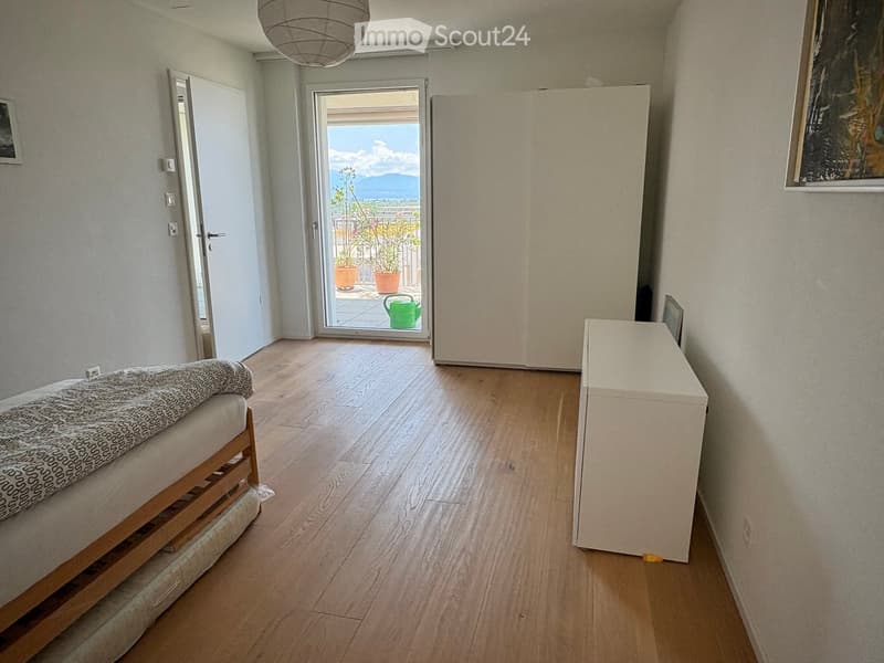 Bright bedroom with wooden flooring, a bed with a patterned blanket, a nightstand, and a view of a balcony with plants.