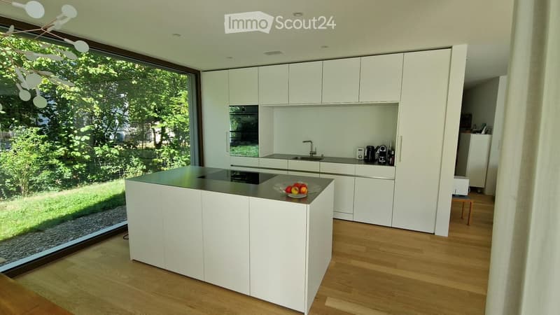 Modern kitchen with white cabinets, central island, and large window overlooking a green area.