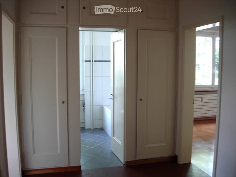 A narrow hallway with wooden flooring, white walls, and several closed doors leading to different rooms. The hallway has a tiled floor in the adjacent bathroom area visible through an open door.