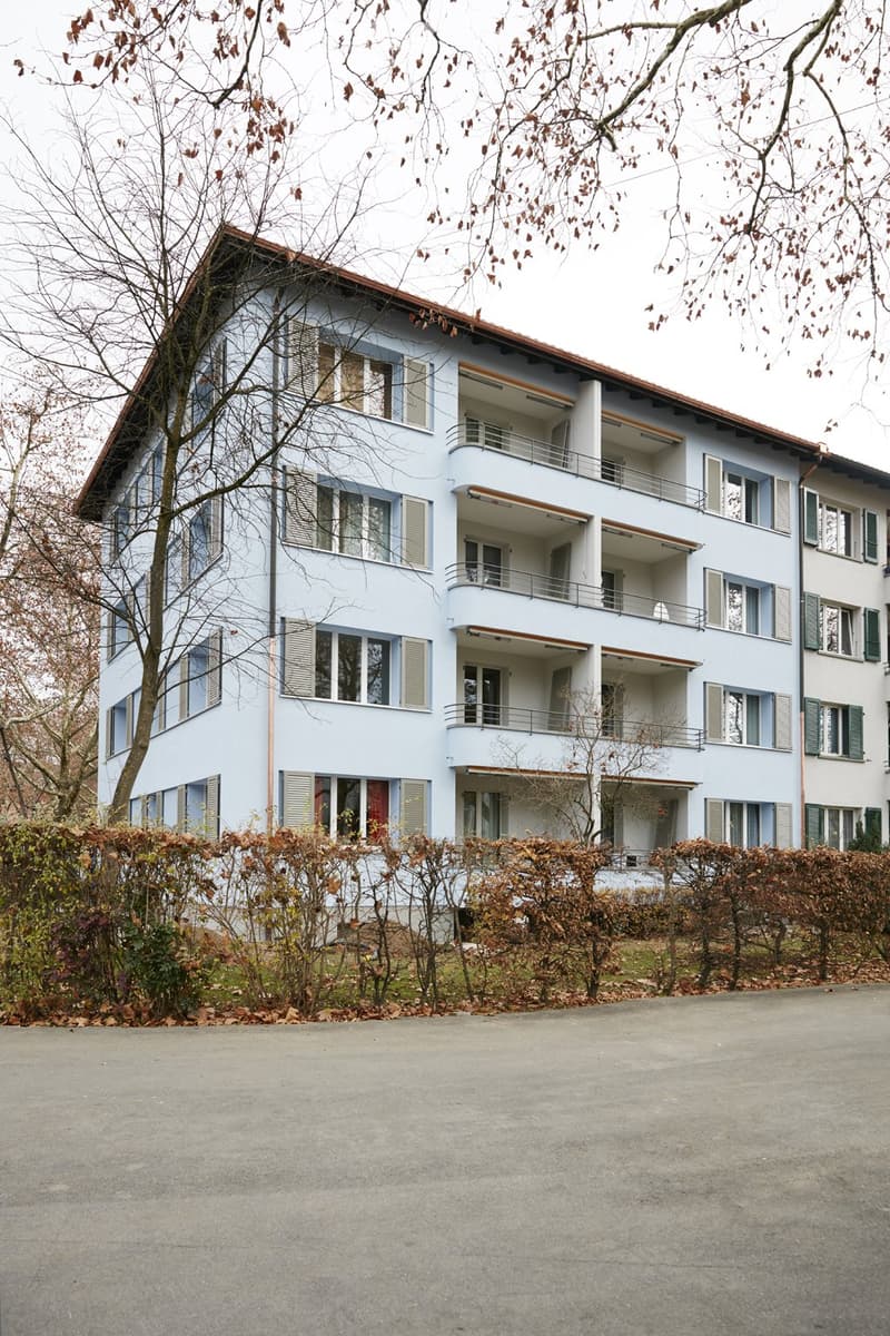 A modern, light blue multi-story building with multiple balconies and large windows, surrounded by bare trees and shrubs.