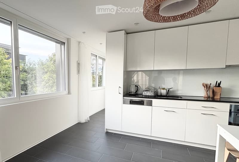 Modern kitchen with white cabinets, dark tiled floor, large windows, and various appliances including a dishwasher and knife block.