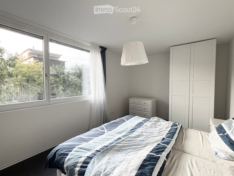 Modern bedroom with a large window, white walls, a bed with blue and white bedding, a wardrobe, a nightstand, and a hanging lamp.