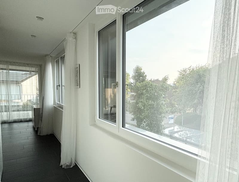 Balcony with large windows, white curtains, and a view of greenery outside.