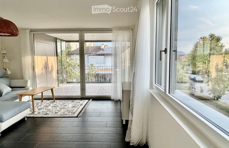 Modern living room with large windows and sliding glass doors leading to a balcony. Features a grey sofa, wooden coffee table, patterned rug, and decorative lamp.